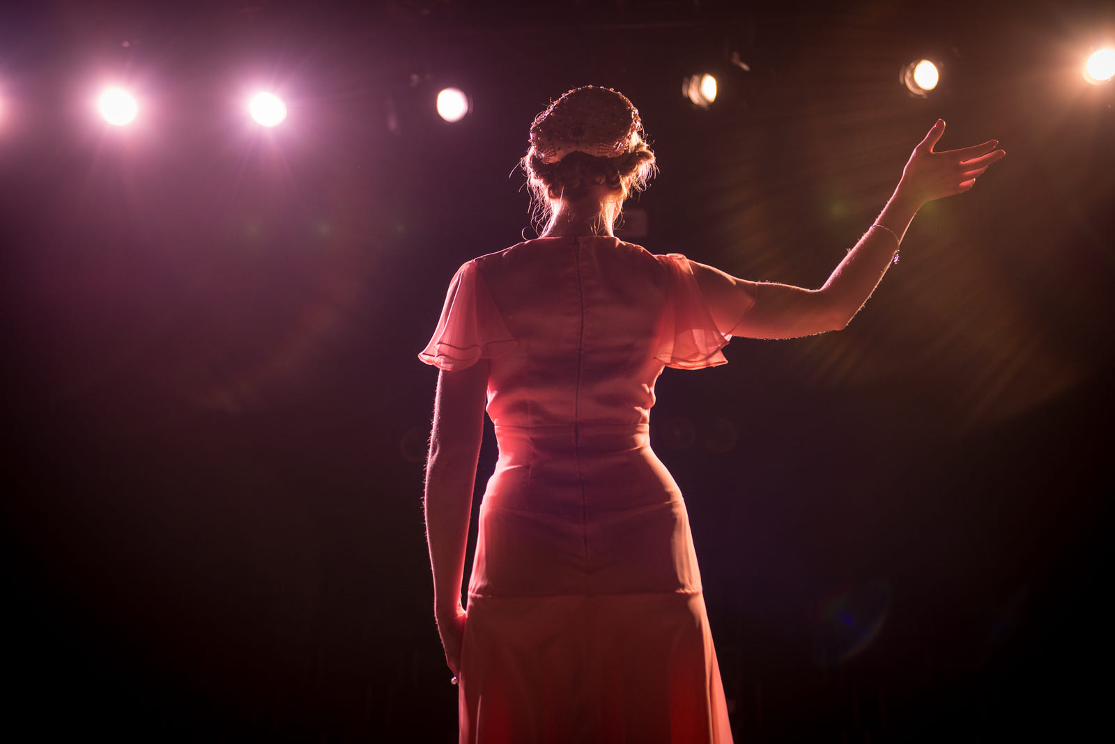 A woman on stage under warm theater lights, arm raised to the room